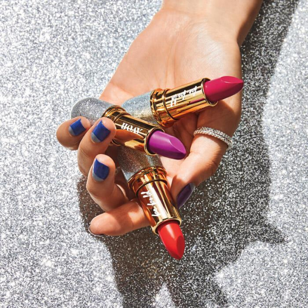 woman's hand holding three tubes of Be Bold Lipstick, in front of a silver sparkling background