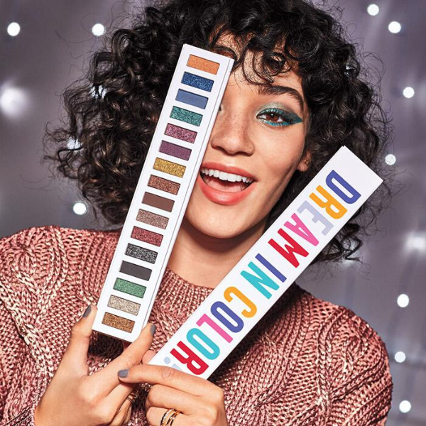 Smiling woman holding a palette of Dream in Color Eye Palette in front of her face, in front of a grey background with hanging lights