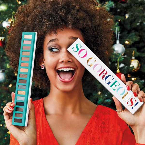 Happy woman holding up the So Gorgeous Eye Palette in front of her face while standing in front of a Christmas tree