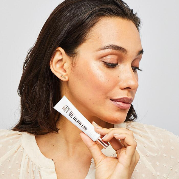 Woman looking towards the lower right corner and holding a tube of Get Me Glowing Dazzle Drops in front of her neck
