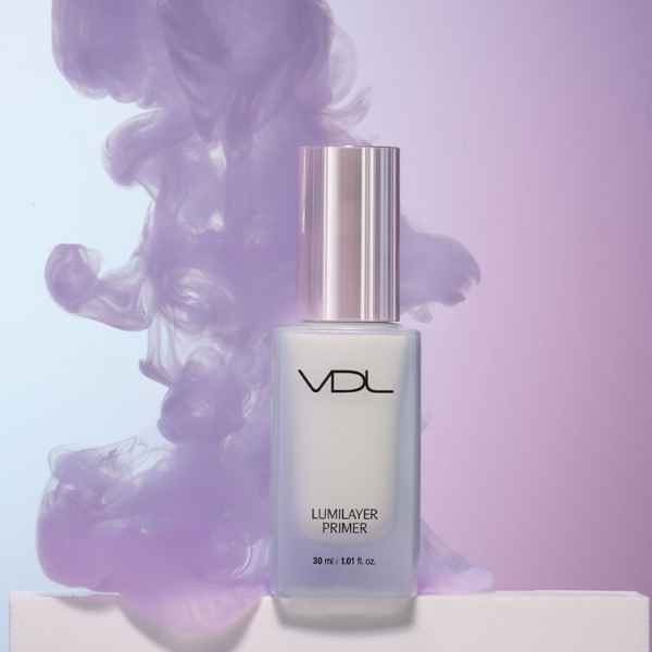 Bottle of Lumilayer primer placed on a white pedestal with an ombre purple and blue background, next to a large puff of light purple smoke
