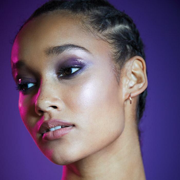 Model with purple eyeshadow and glowing cheeks, looking off to the left while bright pink light reflects off the side of her face, against a purple background