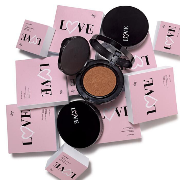 Open compact of Colors of Love Cushion Bronzer on top of a pile of boxes for the product, against a white background