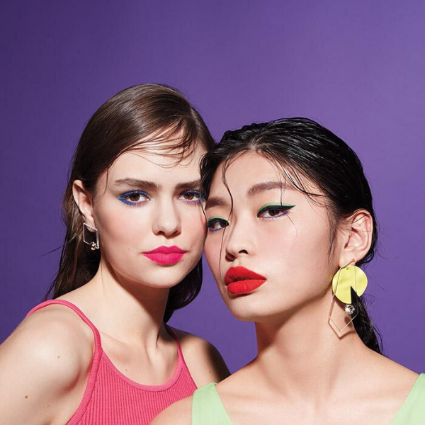 Two women with bright neon makeup standing close together, against a purple background.