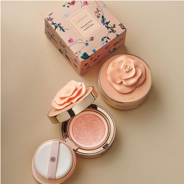 Open compact of Caress Me Cashmere Skin Perfecting Cushion Primer, next to a second closed compact and product box, on top of a beige surface