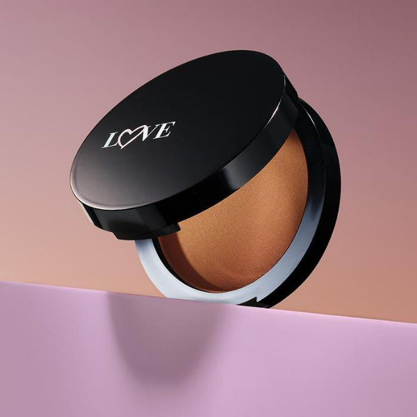 Partially open compact of Warming Bronzer Matte Mineral Powder, sitting on a light purple platform in front of an ombre peach and purple background