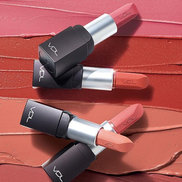 Four open tubes of VDL Expert Real Fit Velvet Lipstick, artistically strewn in front of a striped lipstick smear background