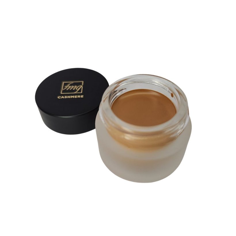 Open frosted glass tub of cream concealer in a medium tan warm shade, next to a black cap that reads "fmg" and "Cashmere", all against a white background